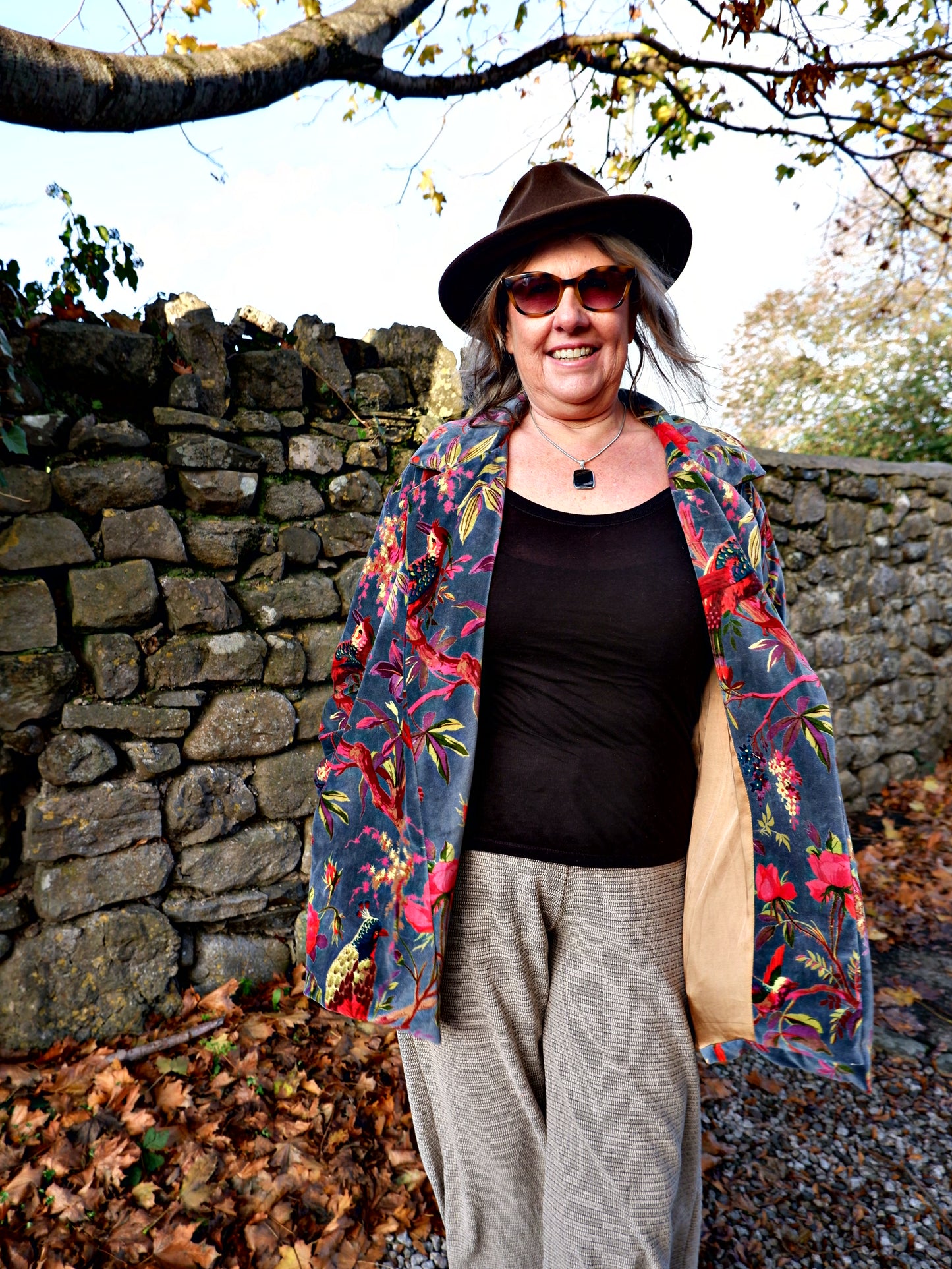 Person wearing a floral-patterned coat over a dark top and hat, standing against a stone wall with trees.