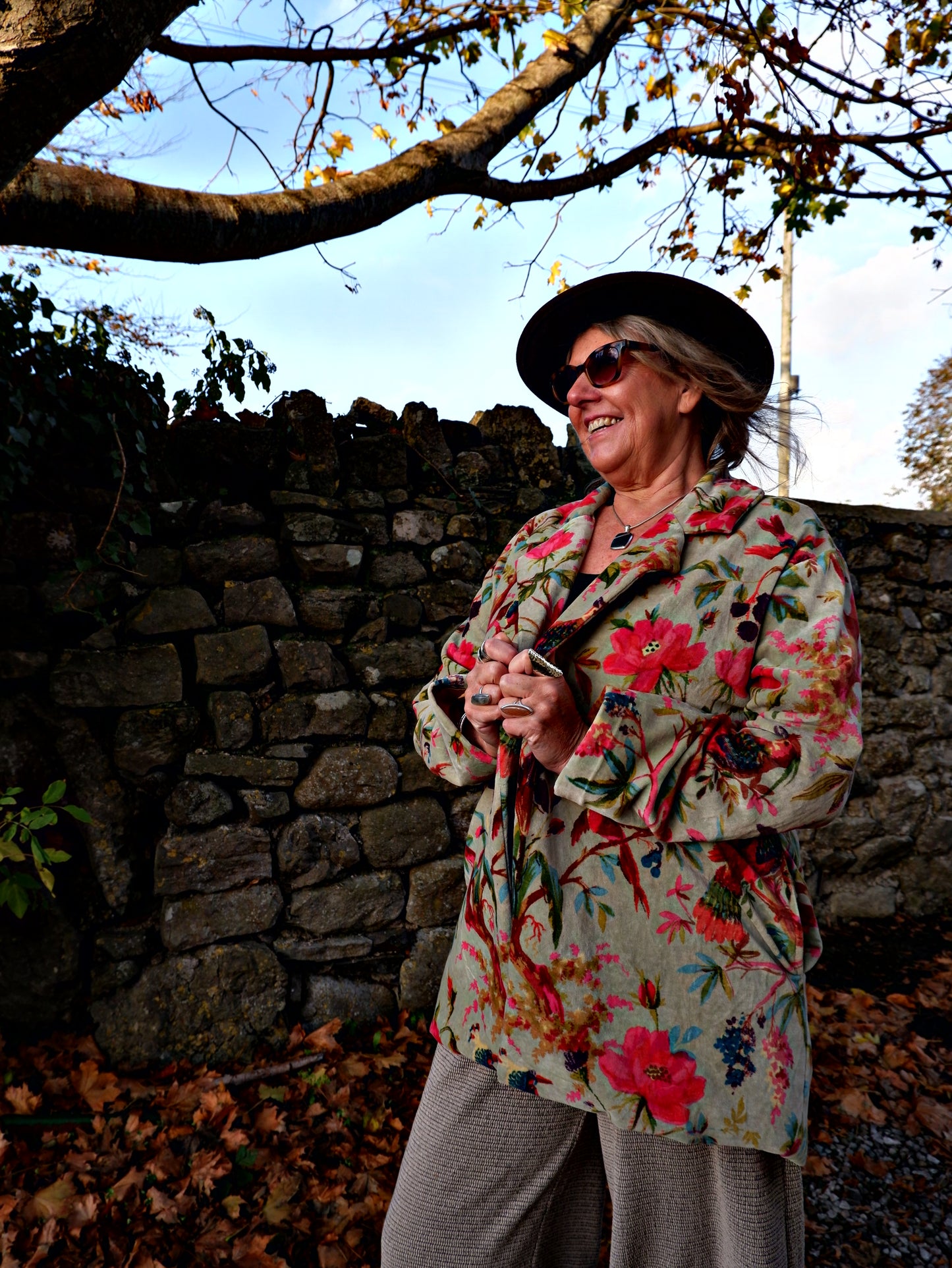 Person wearing a floral coat and hat, standing against a stone wall with trees in the background.