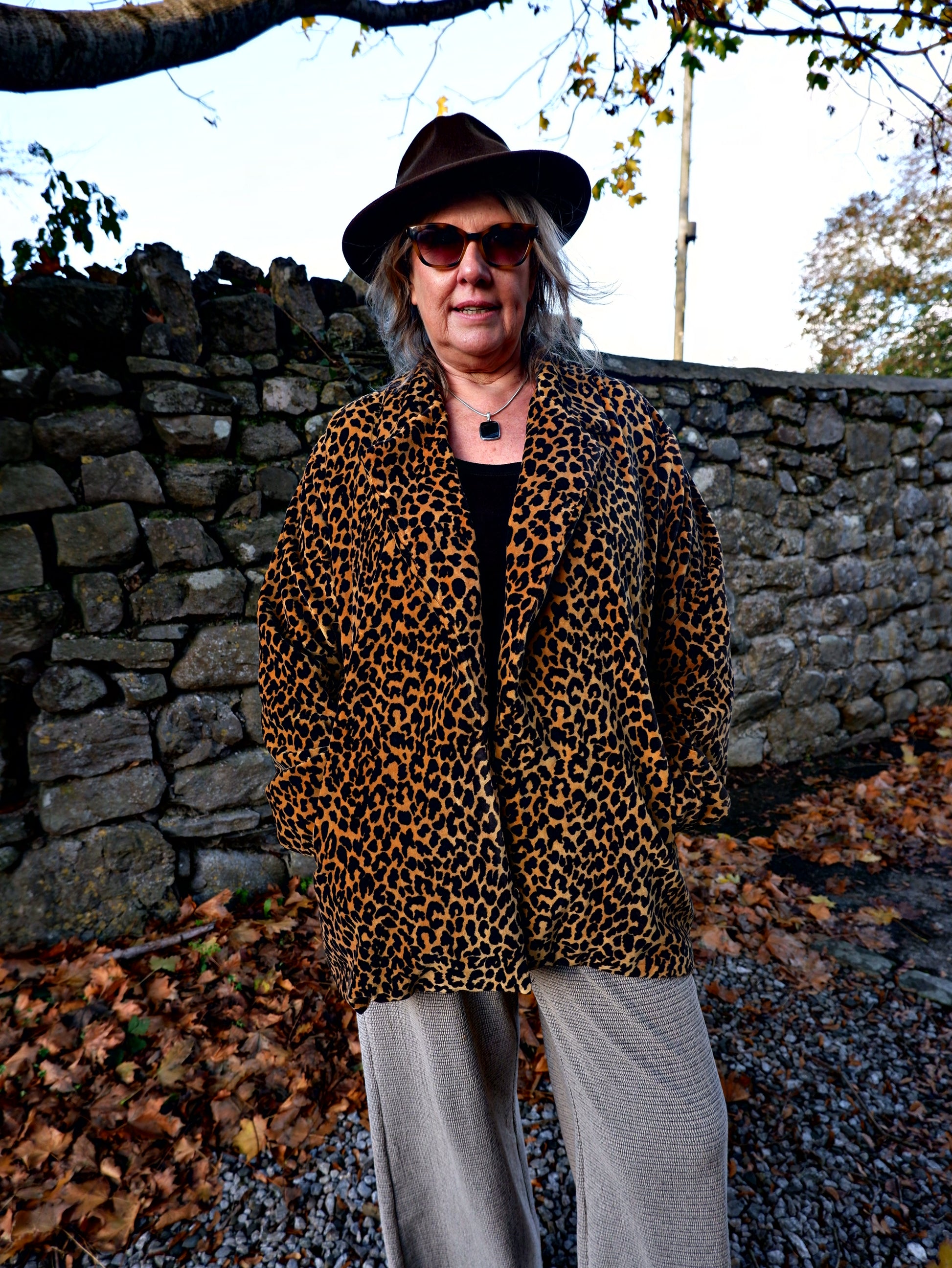 Person wearing a leopard print coat and hat standing on a stone path with autumn leaves.
