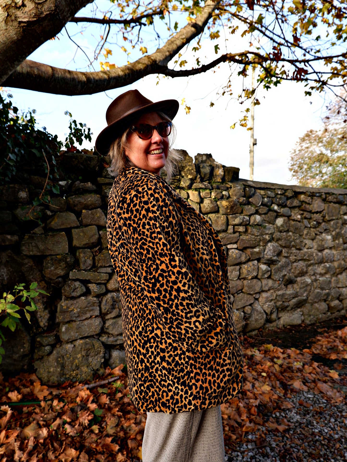 Person wearing a leopard print coat and hat standing near a stone wall with trees in the background