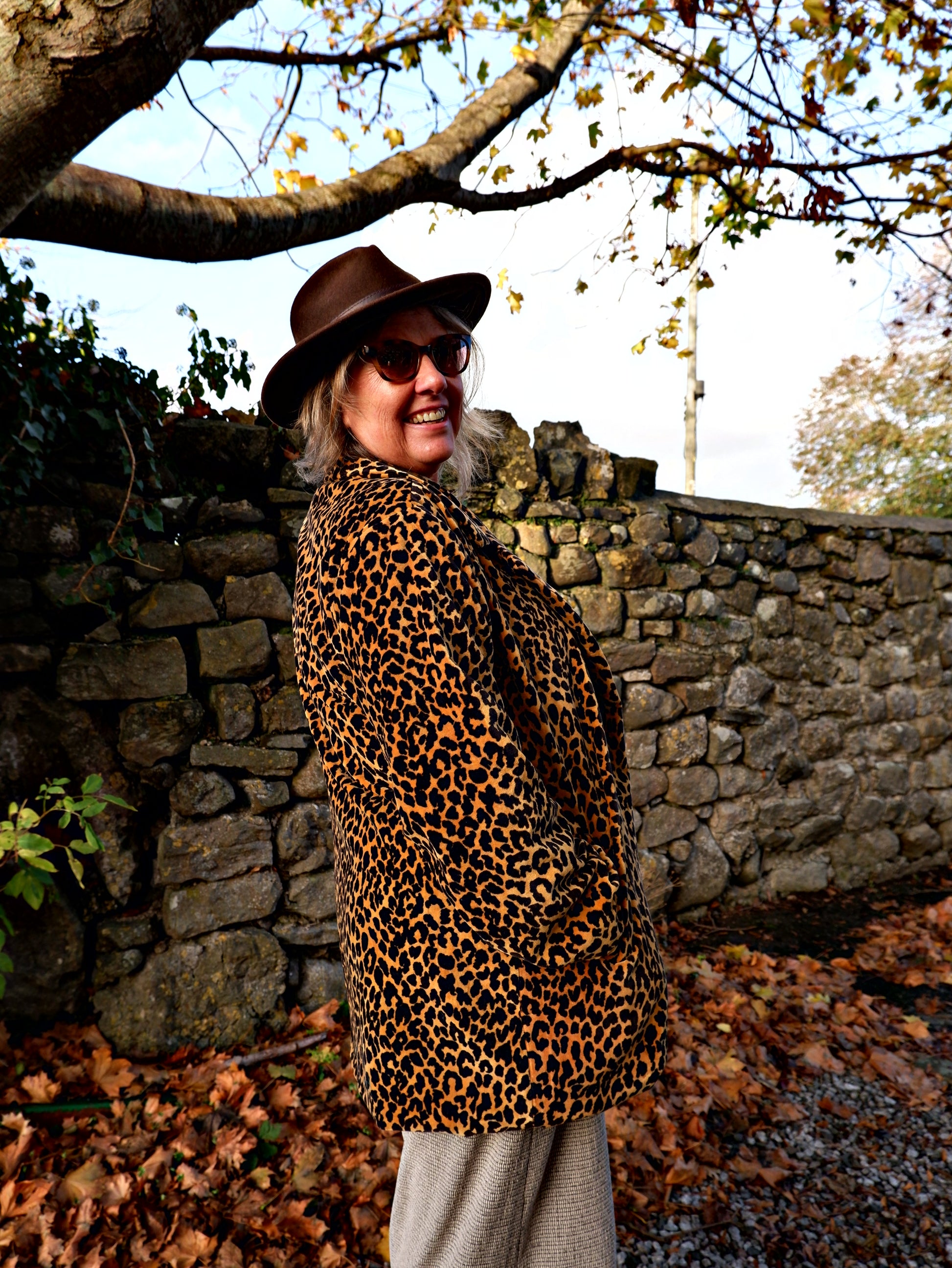 Person wearing a leopard print coat and hat standing near a stone wall with trees in the background