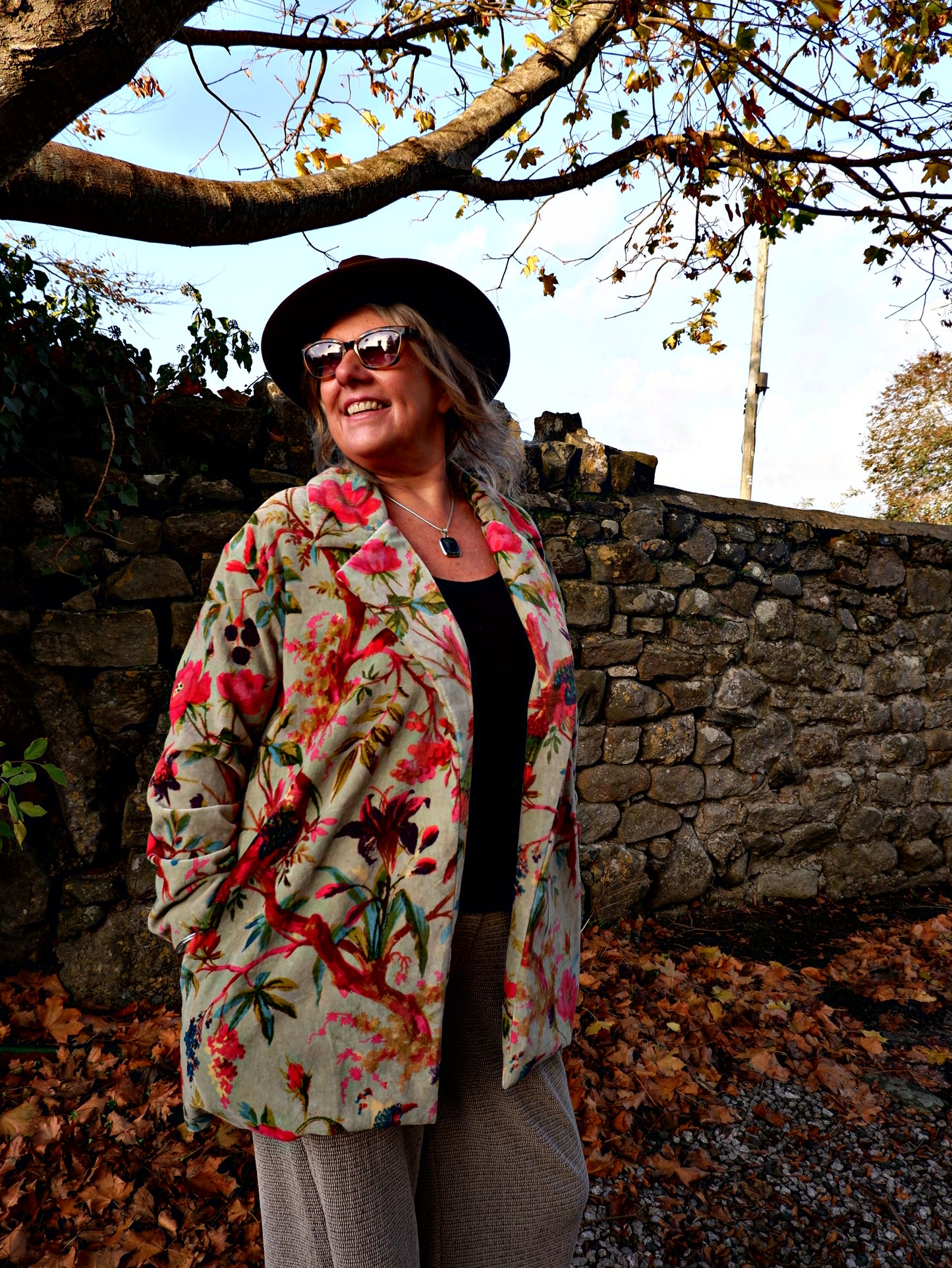 Person wearing a floral coat and hat standing next to a stone wall with trees in the background