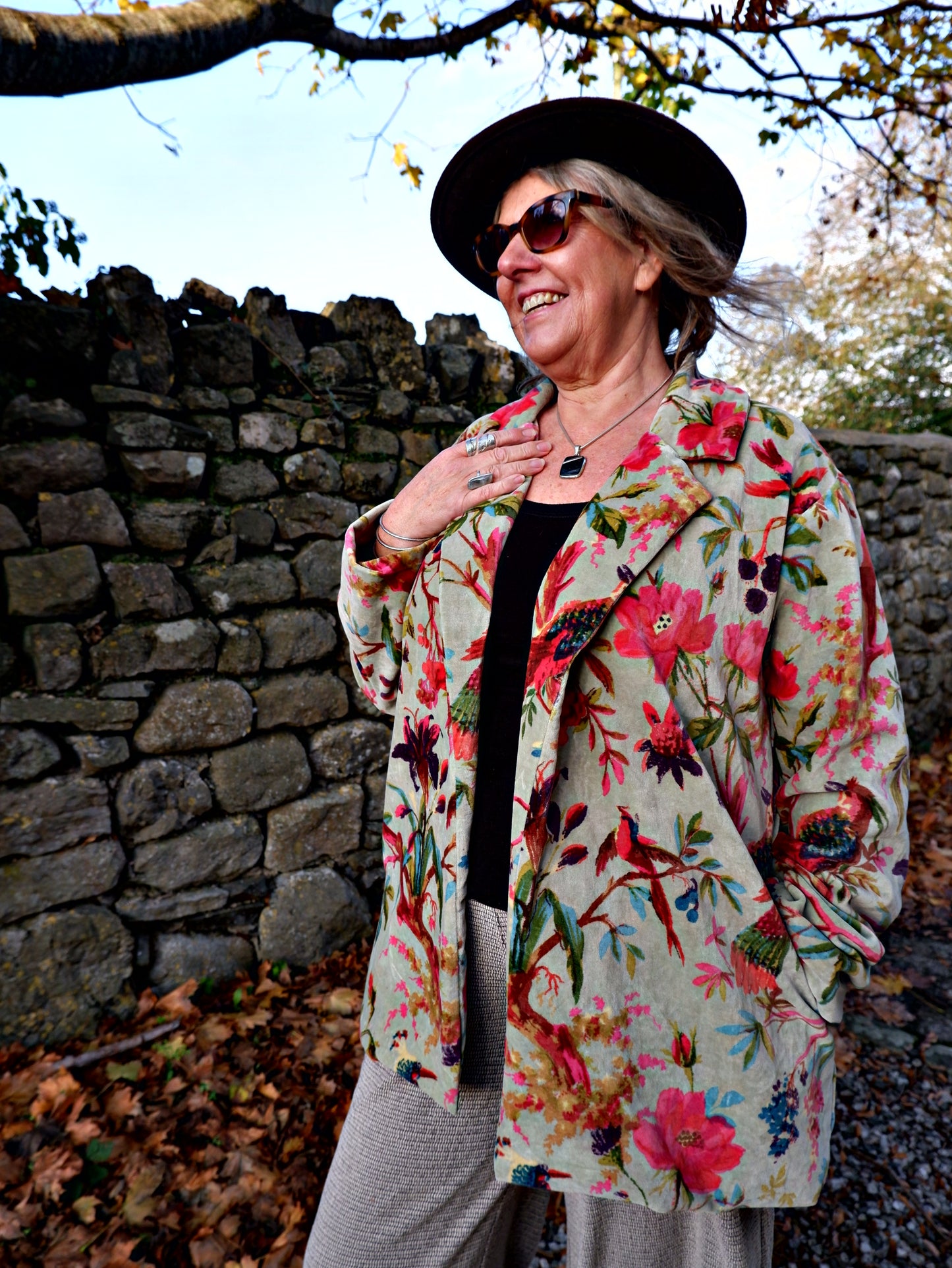 Woman in a floral coat and hat standing against a stone wall with trees in the background